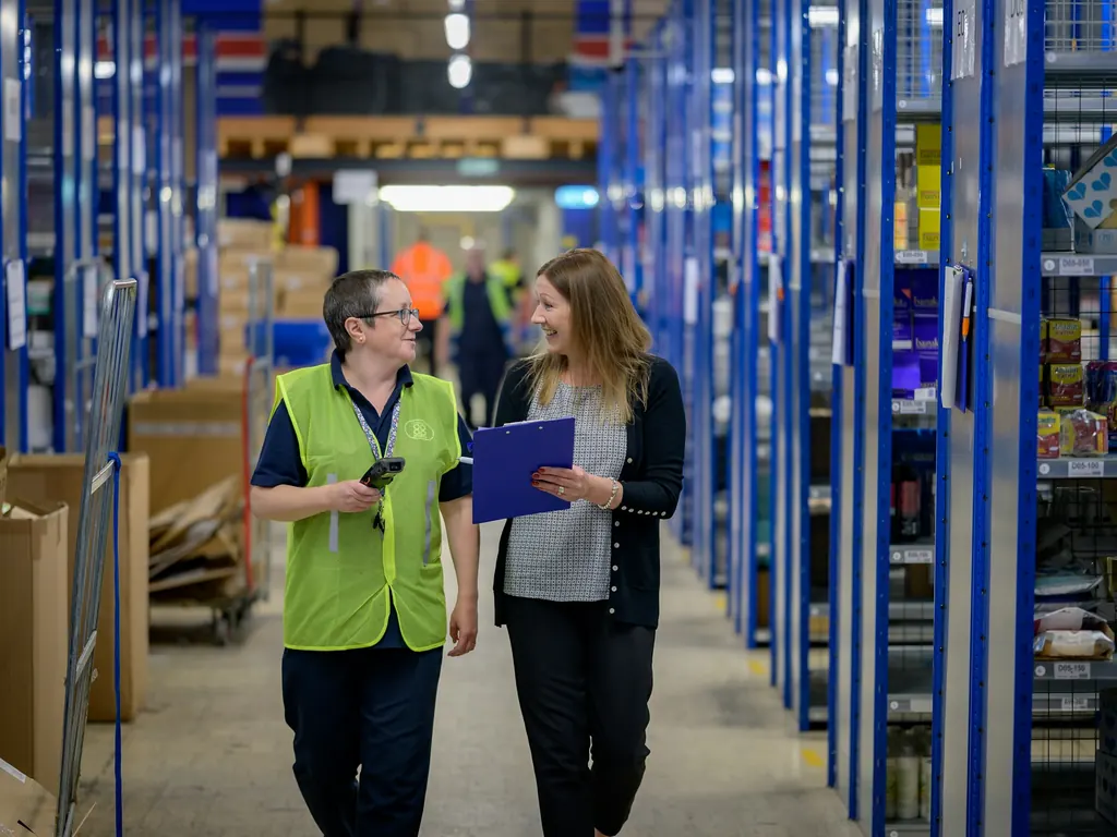 Two women talking as they walk along a warehouse aisle between blue shelving, one in a high visibility vest holding a scanner and the other holding a clipboard