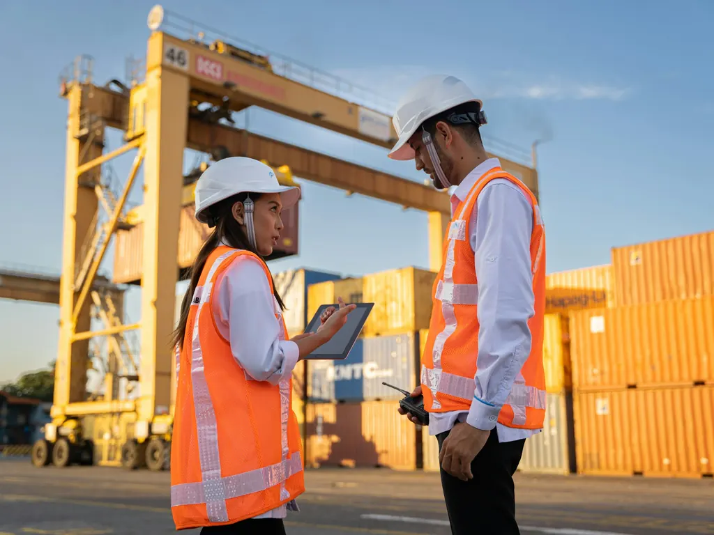 Two workers in white hard hats and high visibility vests talking at a container port, one holding a tablet and the other a radio