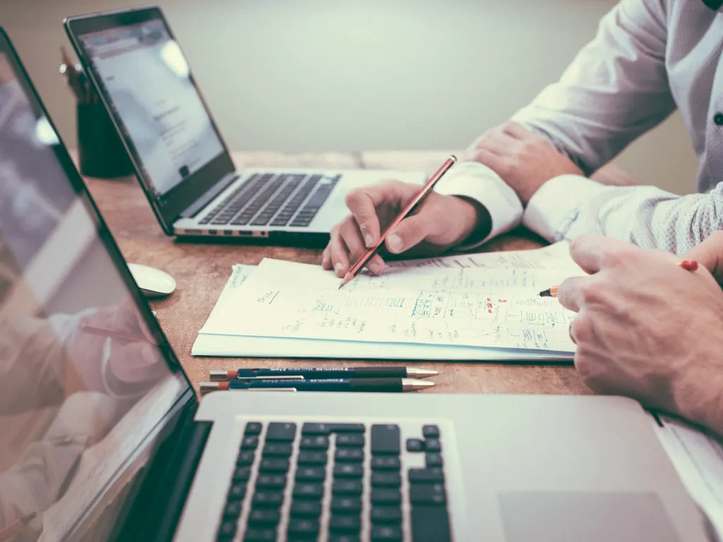 Two people at a table marking up printed pages with a pencil beside two laptops