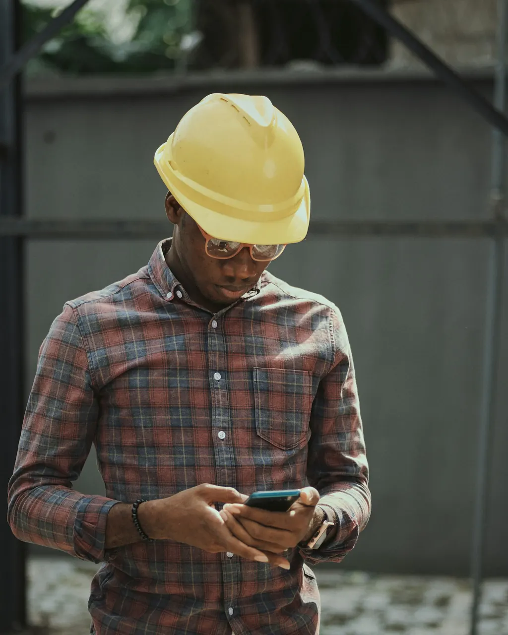 A man in a yellow hard hat, safety glasses and a checked work shirt looking down at a smartphone