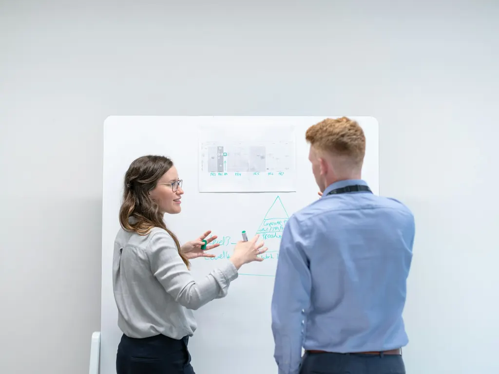 Two people at a whiteboard, one explaining a staged diagram and a pinned chart to the other