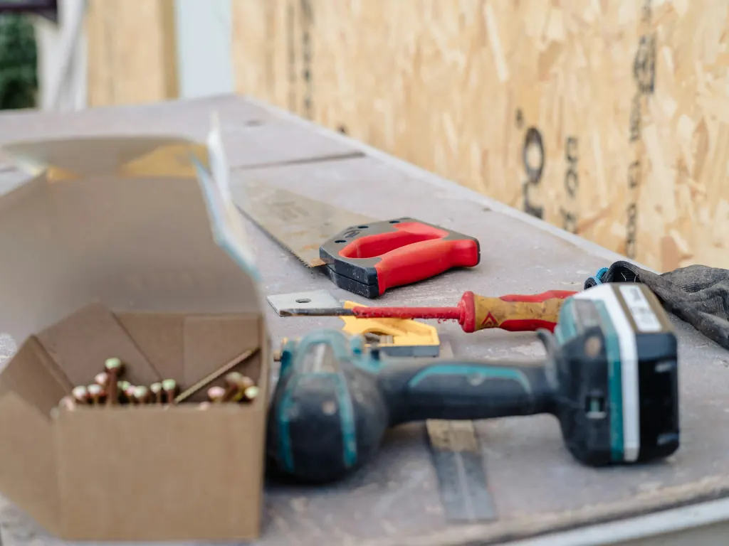 A cordless drill, a handsaw, a screwdriver, work gloves and a box of screws laid out on a site workbench beside sheets of timber