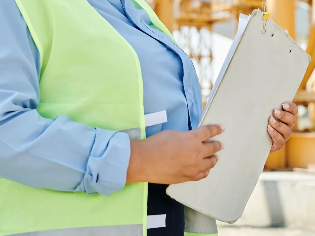 Close up of hands holding a clipboard against a high visibility vest, with yellow scaffolding behind